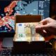 🎨 Portada Sugerida (16:9) Descripción: Un plano detalle de una mano guardando dos billetes de 50 euros dentro de una caja metálica pequeña o un cajón de madera. Al fondo, la pantalla de una computadora muestra un mapa de Europa con zonas de calor. Estética de sobriedad y realismo, con luz de hogar nocturna. Sin texto.