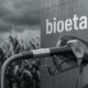 Un primer plano de un pico de manguera de estación de servicio cargando combustible en el tanque de un auto moderno. En el fondo desenfocado, se ve un campo de maíz bajo el sol (materia prima del bioetanol). Estética limpia, industrial y dinámica. Sin texto.