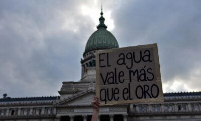Portada Sugerida (16:9) Descripción: Un plano contrapicado de la cúpula del Congreso de la Nación Argentina, con el cielo nublado. En primer plano, un cartel artesanal que dice "El agua vale más que el oro" sostenido por una mano joven. Estética de protesta social con tonos fríos y grises