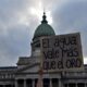 Portada Sugerida (16:9) Descripción: Un plano contrapicado de la cúpula del Congreso de la Nación Argentina, con el cielo nublado. En primer plano, un cartel artesanal que dice "El agua vale más que el oro" sostenido por una mano joven. Estética de protesta social con tonos fríos y grises