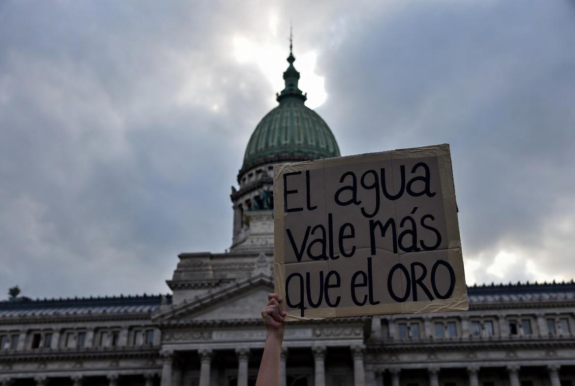 Portada Sugerida (16:9) Descripción: Un plano contrapicado de la cúpula del Congreso de la Nación Argentina, con el cielo nublado. En primer plano, un cartel artesanal que dice "El agua vale más que el oro" sostenido por una mano joven. Estética de protesta social con tonos fríos y grises