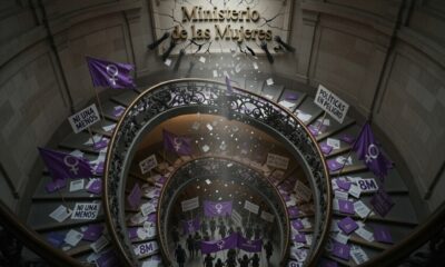 Editorial news illustration about women's rights policies being dismantled in Argentina, symbolic scene: a closed government building with the sign "Ministerio de las Mujeres" being dismantled, purple feminist flags and protest signs in the foreground, women silhouettes marching on March 8, dramatic lighting, newspaper cover style, political illustration, realistic, cinematic, high detail,