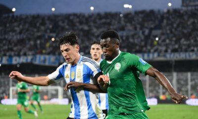 Action sports photography, 4K resolution. Mid-game shot at La Bombonera stadium. An Argentine defender, wearing the official light blue and white striped kit with the three stars over the shield, is in a shoulder-to-shoulder duel for the football against a Mauritanian forward wearing the green national team kit. Their focus is intense. The background shows the blurred texture of the stadium stands filled with cheering fans under modern floodlights at dusk. Fast shutter speed capturing dynamic motion, sweat, and grass kicking up from their boots. Realistic colors and texture.