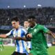Action sports photography, 4K resolution. Mid-game shot at La Bombonera stadium. An Argentine defender, wearing the official light blue and white striped kit with the three stars over the shield, is in a shoulder-to-shoulder duel for the football against a Mauritanian forward wearing the green national team kit. Their focus is intense. The background shows the blurred texture of the stadium stands filled with cheering fans under modern floodlights at dusk. Fast shutter speed capturing dynamic motion, sweat, and grass kicking up from their boots. Realistic colors and texture.