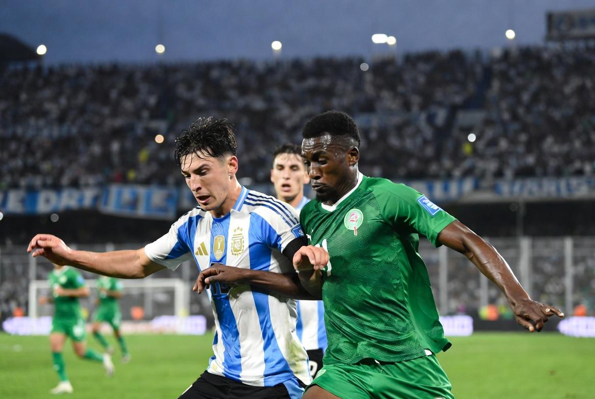 Action sports photography, 4K resolution. Mid-game shot at La Bombonera stadium. An Argentine defender, wearing the official light blue and white striped kit with the three stars over the shield, is in a shoulder-to-shoulder duel for the football against a Mauritanian forward wearing the green national team kit. Their focus is intense. The background shows the blurred texture of the stadium stands filled with cheering fans under modern floodlights at dusk. Fast shutter speed capturing dynamic motion, sweat, and grass kicking up from their boots. Realistic colors and texture.
