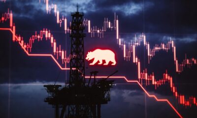 Editorial financial style, 16:9, dark aesthetic, high contrast. A powerful, minimalist silhouette of an offshore oil rig against a deep indigo and crimson digital sky, symbolizing the easing Brent/WTI prices. Faint, downward-trending glowing red data streams representing stock tickers are integrated into the chart, converging towards the lower portion of the frame. A central, prominent digital icon of a 'bear' is integrated into the chart, glowing in a sharp red hue. Professional, clean digital interface look, dark background, no text.