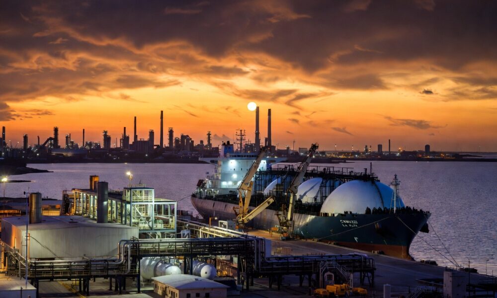 Prompt para Generador de Imágenes (formato 16:9) Descripción Visual: Una fotografía industrial de estilo cinematográfico, con una relación de aspecto de 16:9. La imagen captura la actividad en el Puerto La Plata durante el atardecer, centrada en la operación de un gran buque metanero de GNL y la nueva terminal en Ensenada. Sujetos Principales: Buque Metanero de GNL (Centro-Derecha): Un enorme barco transportador de Gas Natural Licuado (GNL) de doble casco, con sus características cuatro o cinco esferas de almacenamiento gigantes y blancas sobresaliendo de la cubierta. El casco es de color rojo oscuro y negro, con el nombre "CAMUZZI LA PLATA" visible en la proa. Está atracado en un muelle especializado. Terminal de GNL (Centro-Izquierda): En tierra, se extiende una moderna planta de procesamiento y almacenamiento de GNL. Se destacan grandes tanques criogénicos cilíndricos de color plateado y blanco, una compleja red de tuberías amarillas y azules, y brazos de carga articulados conectándose al barco. La estructura tiene un diseño industrial limpio y tecnológico. Escenario y Atmósfera: Puerto La Plata (Fondo): La escena se desarrolla en la desembocadura del Río de la Plata. Al fondo, se aprecia la silueta industrial de Ensenada y Berisso, con chimeneas de la refinería y otras terminales portuarias bajo un cielo dramático de atardecer. Atardecer y Luces: El sol se está poniendo en el horizonte, tiñendo el cielo de tonos naranjas, dorados y violetas profundos, reflejándose intensamente en el agua tranquila del río. La terminal y el barco comienzan a encender sus luces de trabajo LED blancas y amarillas, creando un contraste vibrante con el entorno sombrío.