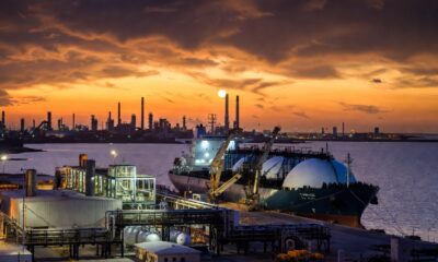 Prompt para Generador de Imágenes (formato 16:9) Descripción Visual: Una fotografía industrial de estilo cinematográfico, con una relación de aspecto de 16:9. La imagen captura la actividad en el Puerto La Plata durante el atardecer, centrada en la operación de un gran buque metanero de GNL y la nueva terminal en Ensenada. Sujetos Principales: Buque Metanero de GNL (Centro-Derecha): Un enorme barco transportador de Gas Natural Licuado (GNL) de doble casco, con sus características cuatro o cinco esferas de almacenamiento gigantes y blancas sobresaliendo de la cubierta. El casco es de color rojo oscuro y negro, con el nombre "CAMUZZI LA PLATA" visible en la proa. Está atracado en un muelle especializado. Terminal de GNL (Centro-Izquierda): En tierra, se extiende una moderna planta de procesamiento y almacenamiento de GNL. Se destacan grandes tanques criogénicos cilíndricos de color plateado y blanco, una compleja red de tuberías amarillas y azules, y brazos de carga articulados conectándose al barco. La estructura tiene un diseño industrial limpio y tecnológico. Escenario y Atmósfera: Puerto La Plata (Fondo): La escena se desarrolla en la desembocadura del Río de la Plata. Al fondo, se aprecia la silueta industrial de Ensenada y Berisso, con chimeneas de la refinería y otras terminales portuarias bajo un cielo dramático de atardecer. Atardecer y Luces: El sol se está poniendo en el horizonte, tiñendo el cielo de tonos naranjas, dorados y violetas profundos, reflejándose intensamente en el agua tranquila del río. La terminal y el barco comienzan a encender sus luces de trabajo LED blancas y amarillas, creando un contraste vibrante con el entorno sombrío.