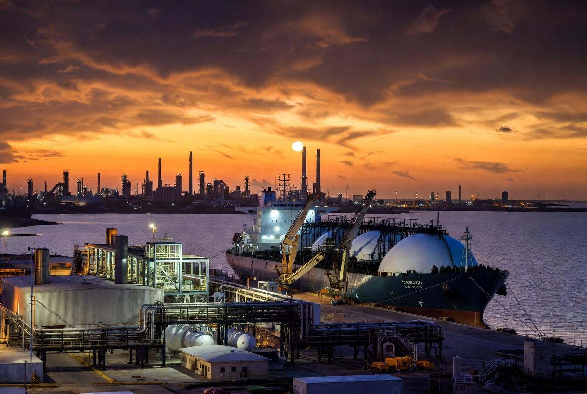 Prompt para Generador de Imágenes (formato 16:9) Descripción Visual: Una fotografía industrial de estilo cinematográfico, con una relación de aspecto de 16:9. La imagen captura la actividad en el Puerto La Plata durante el atardecer, centrada en la operación de un gran buque metanero de GNL y la nueva terminal en Ensenada. Sujetos Principales: Buque Metanero de GNL (Centro-Derecha): Un enorme barco transportador de Gas Natural Licuado (GNL) de doble casco, con sus características cuatro o cinco esferas de almacenamiento gigantes y blancas sobresaliendo de la cubierta. El casco es de color rojo oscuro y negro, con el nombre "CAMUZZI LA PLATA" visible en la proa. Está atracado en un muelle especializado. Terminal de GNL (Centro-Izquierda): En tierra, se extiende una moderna planta de procesamiento y almacenamiento de GNL. Se destacan grandes tanques criogénicos cilíndricos de color plateado y blanco, una compleja red de tuberías amarillas y azules, y brazos de carga articulados conectándose al barco. La estructura tiene un diseño industrial limpio y tecnológico. Escenario y Atmósfera: Puerto La Plata (Fondo): La escena se desarrolla en la desembocadura del Río de la Plata. Al fondo, se aprecia la silueta industrial de Ensenada y Berisso, con chimeneas de la refinería y otras terminales portuarias bajo un cielo dramático de atardecer. Atardecer y Luces: El sol se está poniendo en el horizonte, tiñendo el cielo de tonos naranjas, dorados y violetas profundos, reflejándose intensamente en el agua tranquila del río. La terminal y el barco comienzan a encender sus luces de trabajo LED blancas y amarillas, creando un contraste vibrante con el entorno sombrío.